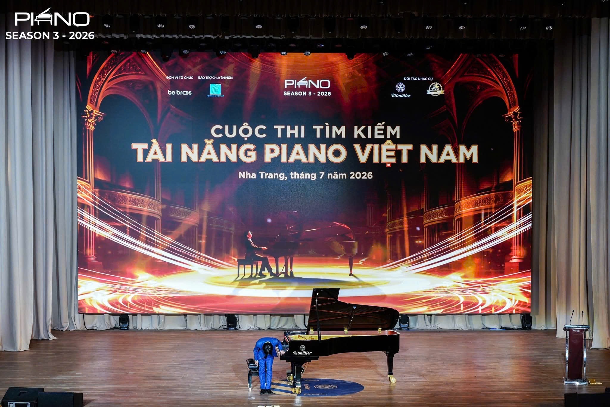 Sân khấu Cuộc thi Tìm kiếm Tài năng Piano Việt Nam 2026 tại Nha Trang với màn hình LED lớn in tên cuộc thi và dòng chữ Nha Trang, tháng 7 năm 2026; trên sàn gỗ là cây grand piano Ritmüller, hai bên là rèm sân khấu