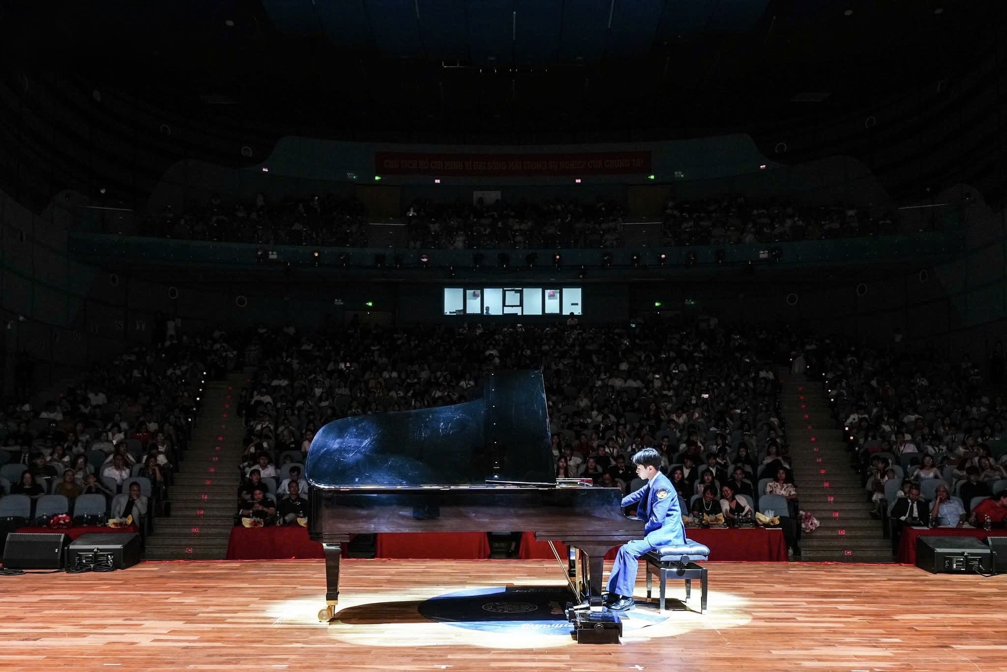 Toàn cảnh một khán phòng lớn kín khán giả, cây grand piano đen đặt giữa sân khấu gỗ dưới luồng đèn spotlight, người biểu diễn mặc bộ đồng phục xanh ngồi bên đàn