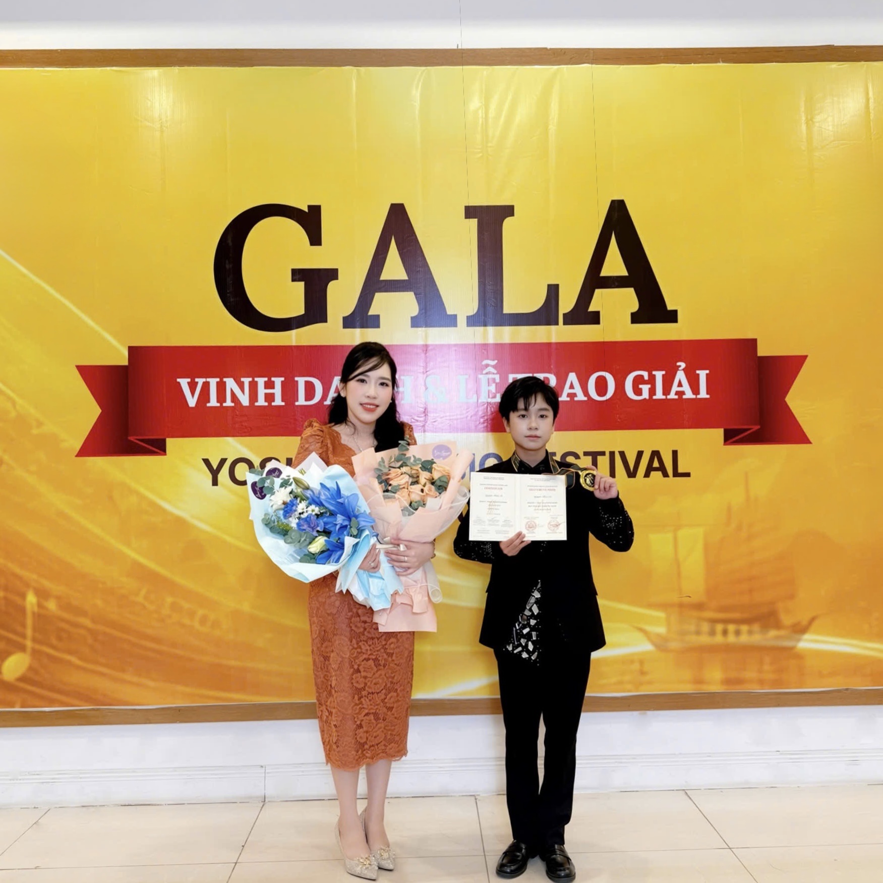 Cô Phù Mai Hiền, giáo viên của Nguyễn Thái An, trong bộ vest đen bên tấm chứng nhận và huy chương, cạnh người đồng hành giữ hai bó hoa, trước phông Gala Vinh danh & Lễ trao giải